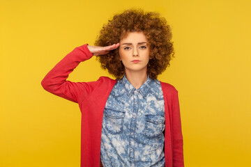 Yes sir! Portrait of serious responsible woman with curly hair giving salute, attentively listening to order and following discipline, obedient behavior. studio shot isolated on yellow background