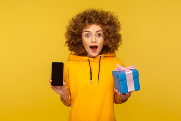 Wow, crazy shopping, buying presents online! Portrait of surprised curly-haired woman in urban style hoodie holding cellphone and gift box, looking with amazement. indoor studio shot yellow background