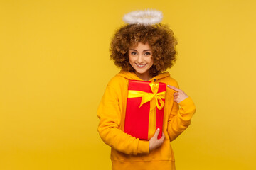 Holiday surprise. Portrait of kind curly-haired angelic woman with saint nimbus pointing at wrapped...