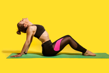 Fitness woman with hair bun in tight sportswear sitting on gym mat doing sport, bending backwards stretching spinal muscles, warming up with flexibility exercises. studio shot, isolated on yellow