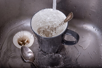 Water flows into a cup with a spoon that stands in a metal sink in the kitchen, top view, close-up