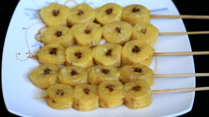 Banana satay on white plate, black background.