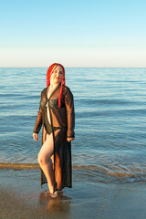 A young woman in a swimsuit and a black cardigan with African pigtails walks along the seashore.