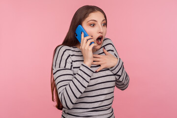 Mobile communication. Portrait of amazed young woman in striped sweatshirt expressing great surprise while talking on cell phone, shocked by unbelievable news. studio shot isolated on pink background