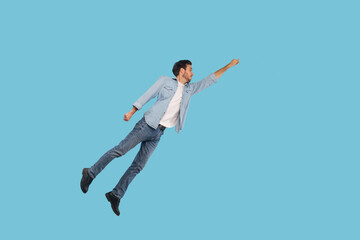 Full length portrait of man in denim outfit flying up as superhero with raised hand and inspired ambitious expression, achieving goal, jumping high in air. studio shot isolated on blue background