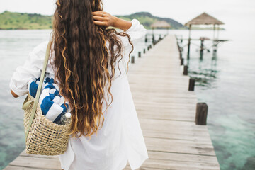 Woman with handmade wicker bag, two beach towels and glass bottle for water going to the beach. View from behind close up. Eco friendly and zero waste concept.