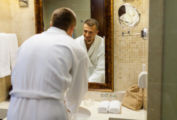 The guy smiles in a white coat and looks at himself in the bathroom mirror in the morning. Starting a new day with a smile. Handsome young man standing in front of a mirror