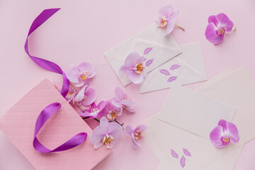 Pink gift bag and flying orchid flowers on light pink background. Top view greeting card with delicate flowers.
