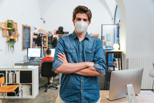 Portrait of businessman in office with arms crossed near work station with medical mask in protection from contamination by Coronavirus, Covid-19 - Millennial man at working for his start up company