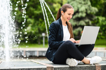 Female student sits near city fountain in the park and is using laptop for preparing homework. An...