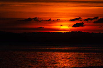 Magical & dramatic scenic landscape shot of sun setting down, only half visible, behind a silhouette of mountains & clouds and the sunshine sparkling in the water. A stunning orange & black scenery.