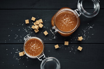 Two glass jars with homemade salted caramel and pieces of brown sugar with salt crystals