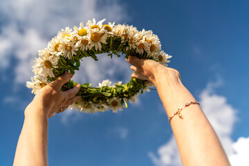 A wreath of white daisies in a woman's hand against a background of blue sky and white clouds © svetograph