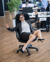 Cheerful woman in a skirt is riding on an office chair at the workplace while colleagues are working. Business lady having fun at work break.