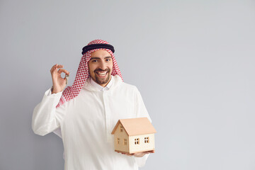Arabian man realtor with wooden house model in his hand over grey background.