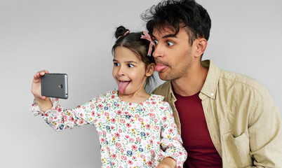 Funny young father and his pretty daughter making faces while taking self portrait on smart phone at home. Dad and cute little girl taking a self-portrait on mobile phone.
