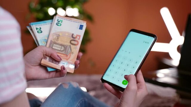 Annual financial report, debit and credit comparison. Female accountant holding stack of 20 and 50 euro banknotes and mobile phone with calculator, woman calculating monthly income of her family or