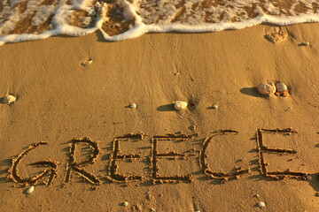 Greece Hand Written on Sandy Texture at a Sunny Beach with Pebbles and the Sea Water in the Background. Travel in the Summer Months Tourism Wallpaper Image. Seascape Vacation in the Greek Islands.