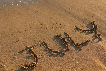 July Hand Written on Sandy Texture at a Sunny Beach with Pebbles with Sea Water in the Background. Travel in the Summer Months Tourism Wallpaper Image. Seascape Vacation Concept.