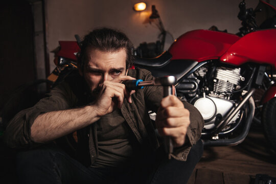 A young man sits in a garage with a motorcycle. A man examines a tool