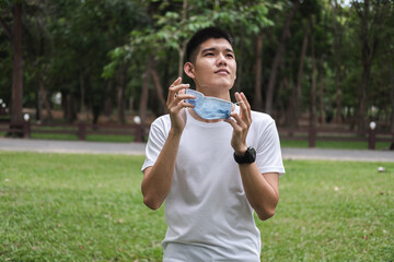 asian young man remove mask from his face while no people for social distancing in park in summer....