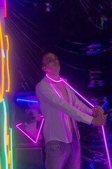 Portrait of the Young Handsome emotional man in an unbuttoned white shirt with flexible pink neon ribbon. Neon Room. Old 90's retro style. Reflect of neon words near male. Selective focus.