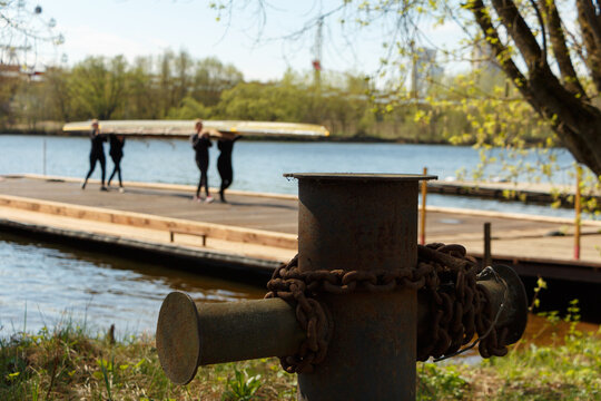 A metal bollard with a rusty chain on the bank of the river against the background of a blurred image of a female rowing crew. 4 girls go on pantone and carry the boat.
