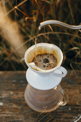 Hand pouring hot water from steel kettle in glass flask dripper with filter on background of rural countryside, herbs in sunset. Alternative coffee brewing. Atmospheric rustic moment. Vertical image