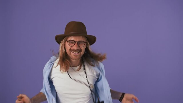 Stylish fellow in casual outfit, hat, glasses and bijouterie. He smiling and dancing while posing against purple studio background. Close up
