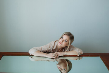 A beautiful girl with light hair is sitting next to a mirror in a room with a cold shade. Fashion shoot for a women's clothing brand