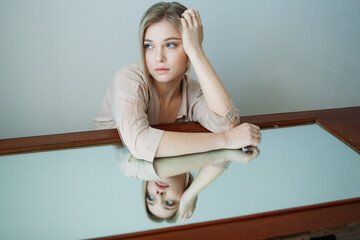 A beautiful girl with light hair is sitting next to a mirror in a room with a cold shade. Fashion shoot for a women's clothing brand