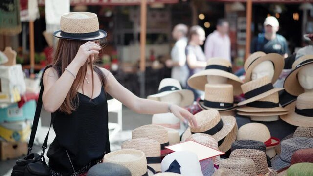 Stylish girl trying on panama hat on local craft market and looking into the mirror. Market counter full of different types and styles of head ware, colorful assortment attracting tourists. Summer