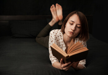 the girl lies with a book in a black bed and carefully reads