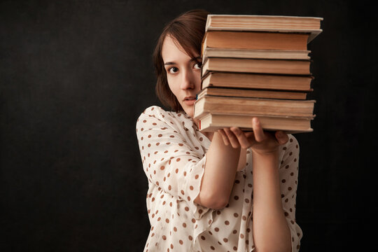 A Young Girl In A Blouse Holds A Lot Of Books. Girl On A Black Background. Girl Peeks Out From Behind Books.