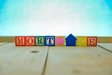 Conceptual of mortgage and home loan. Wooden block letters and  house-shape key chain hand held spelling the words MORTGAGE on wooden table. Focus at blue letter T. Bluish background.
