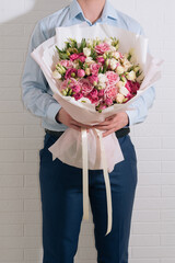 young man in a blue classic shirt and suit holds in his hands a huge bouquet of white-pink roses and eustoma in pink packaging on the white wall background