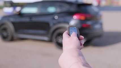 Woman hand presses on the remote control car alarm systems. Female holding car keys with black car on background.