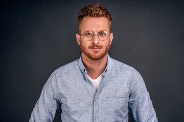Good-looking creative and smart redhead male entrepreneur hiring new employees, standing in casual shirt and glasses over gray wall, smiling friendly, gazing at camera with thoughtful, determined look