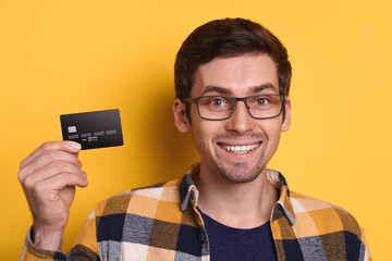 Portrait of happy smiling guy in glasses wearing casual clothing holding credit card, smiling happily isolated on yellow background. Online shopping, ecommerce, NFC, technology and reaction concept