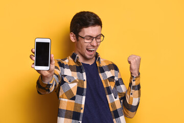 Exited successful man in glasses showing smartphone with blank space and making yes hand gesture isolated on yellow studio background, happy wining online, feel overjoyed, joyful reading good news