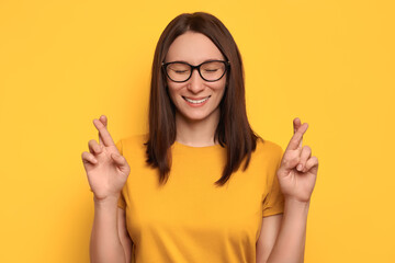 Close-up portrait of hoping beautiful woman crossing her fingers, eyes closed, asking better, smiling, wishing good luck, isolated on yellow wall background. Emotion, feeling, believe and hope concept