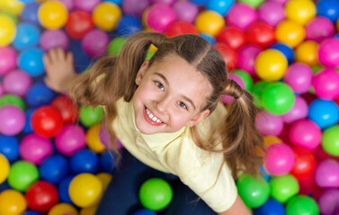 Fototapeta na wymiar Happy girl with ponytails playing in ball pit at kids playground, overhead view