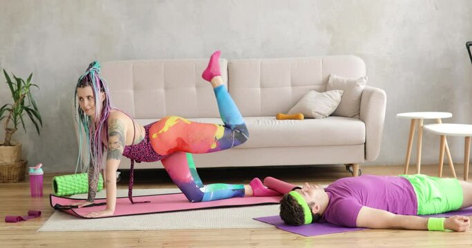 Woman doing sport exercise for legs on mat, man lying on mat near her, fitness at home together. Joke, mem, humor, parody, joking behavior. Workout, training of couple in colourful sportswear.