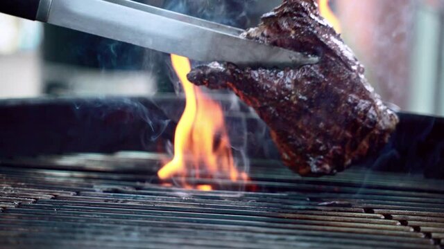 Tasty juicy meat steak roasting on barbecue grill, burning fire and smoke concept. Man using metal cooking tongs for turning aged prime rare steak on bbq grid. BBG party on open terrace