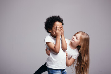 The dark-skinned boy closed his eyes and considers to start playing hide and seek with a fair-haired girl on an gray background.