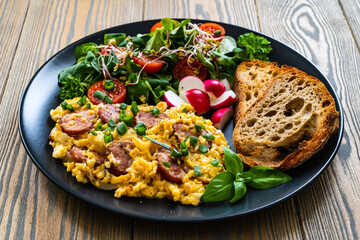 Delicious breakfast - scrambled eggs with fried sausages,  vegetable salad and toasted bread served on black plate on wooden table 
