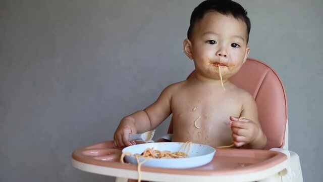 Asian cute little boy sitting and eatting spaghetti on high baby feeding chair and making mess.
