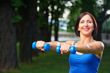 Portrait of cheerful aged woman in fitness wear exercising with dumbbells in park.