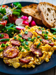 Delicious breakfast - scrambled eggs with fried sausages,  vegetable salad and toasted bread served on black plate on wooden table 
