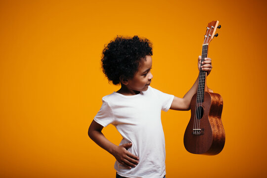 Black-skinned Maracanian boy holds a ukulele guitar in his hand and looks with his hand on his side against an orange background